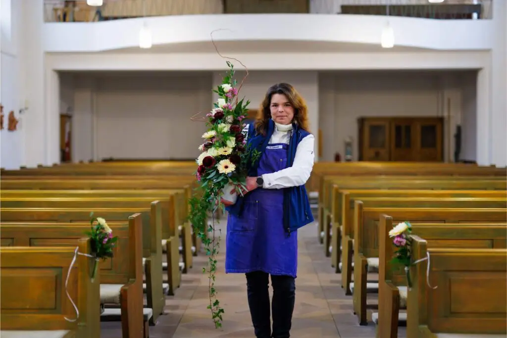 Floristin in Kirche h&auml;lt langes h&auml;ngendes Blumengesteck, B&auml;nke mit Blumen f&uuml;r Hochzeitsdeko