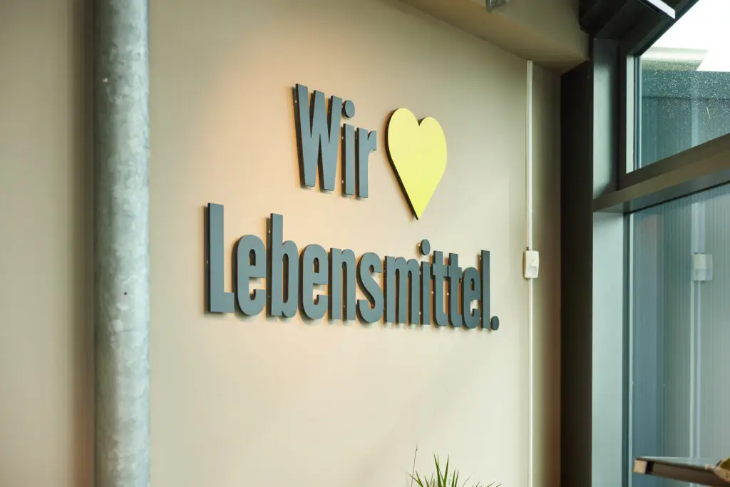 Wandlogo Wir &hearts; Lebensmittel mit gelbem Herz an Innenwand neben Fenster, modernes Supermarkt‑Interieur
