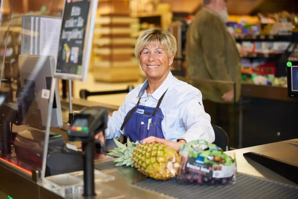 Lächelnde Kassiererin an Supermarktkasse mit Ananas und Trauben auf dem Förderband
