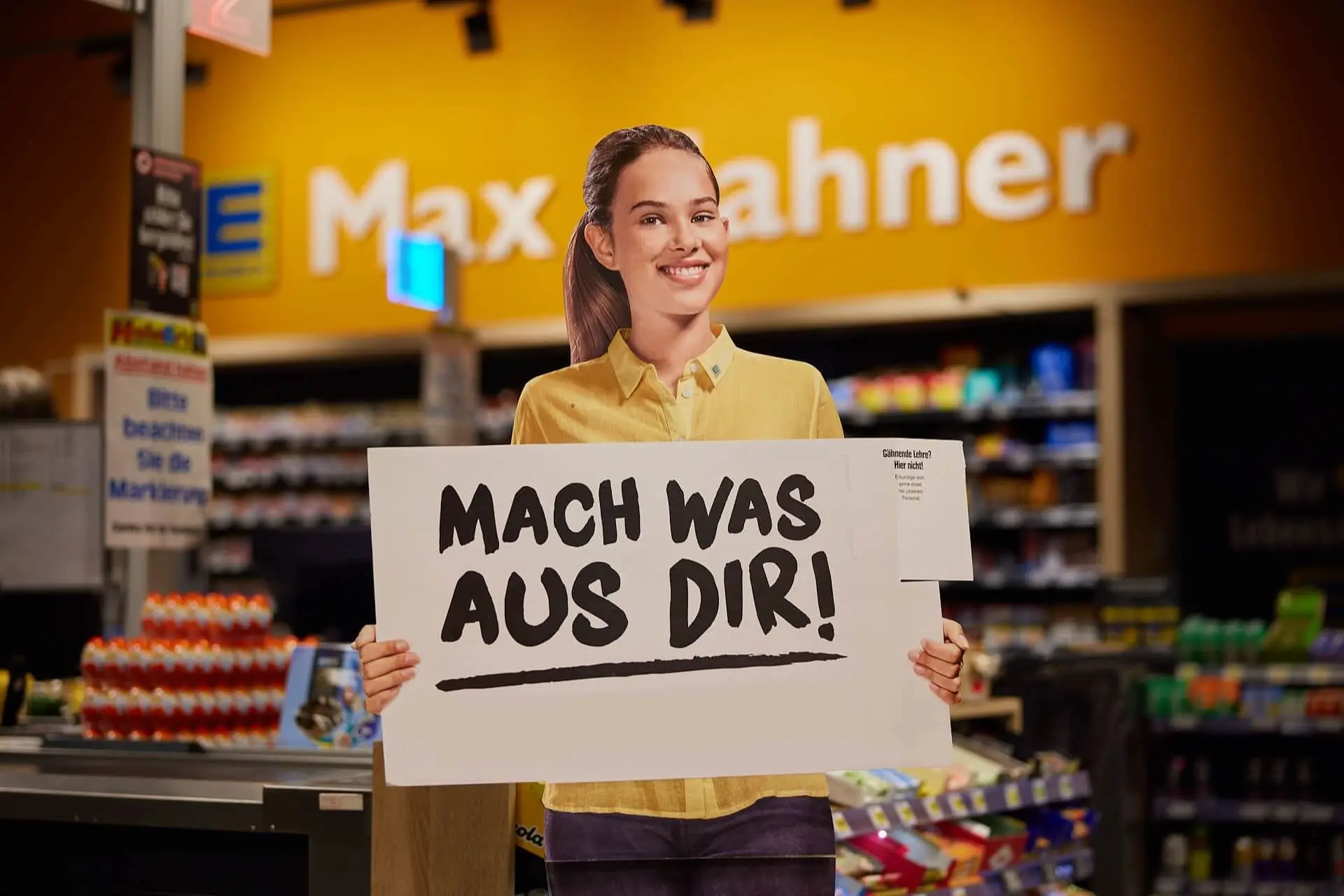 Werbeaufsteller im Supermarkt: l&auml;chelnde Mitarbeiterin h&auml;lt Schild Mach was aus dir! zur Personalwerbung