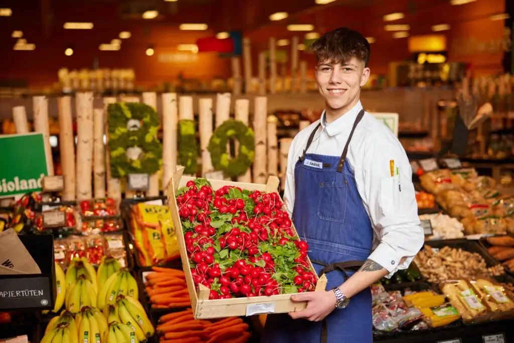 Frische Radieschen im Supermarkt: junger Mitarbeiter h&auml;lt Kiste in der Gem&uuml;seabteilung (Bio-Angebot)