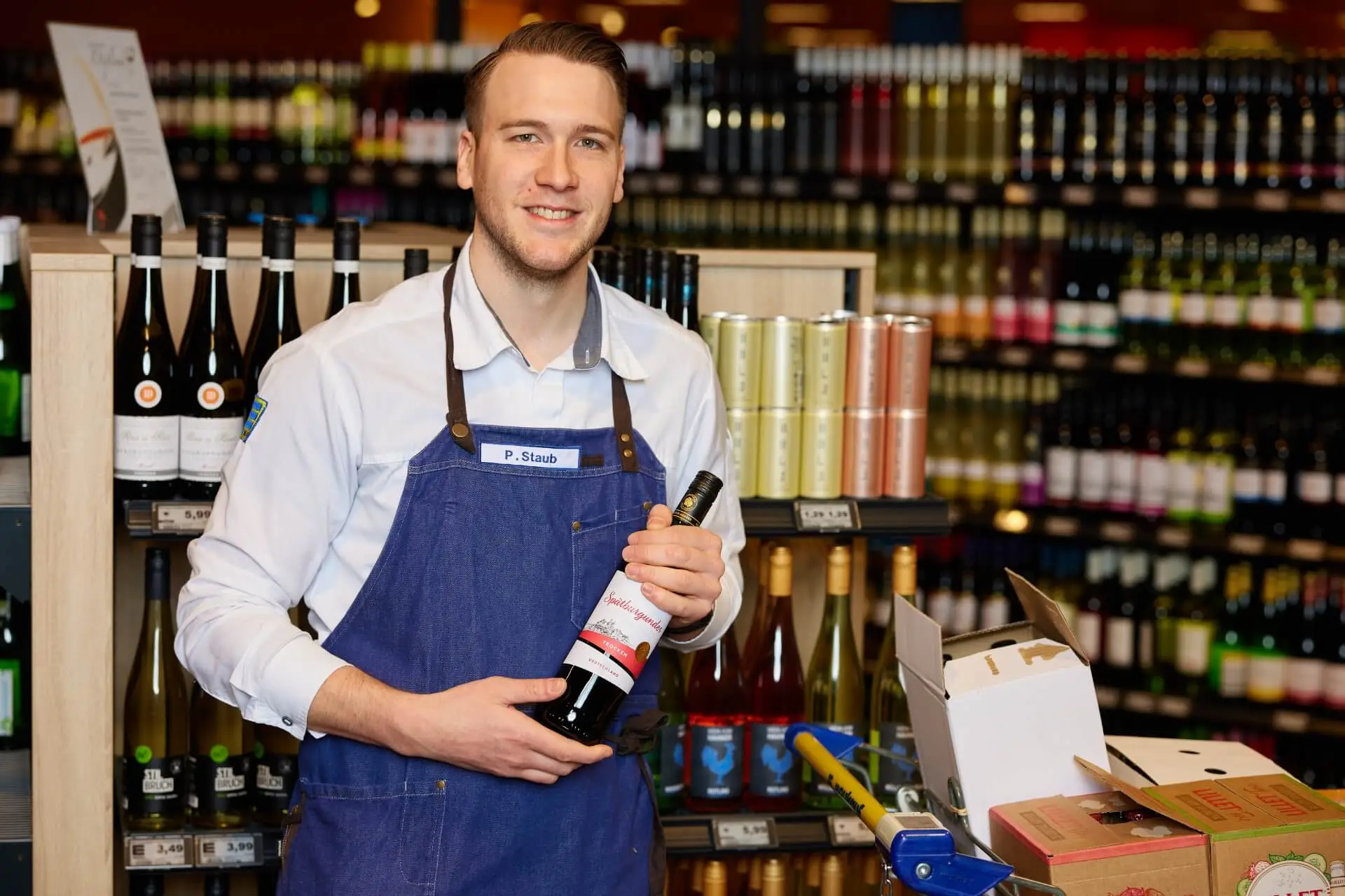Mitarbeiter im Supermarkt h&auml;lt Weinflasche in der Weinabteilung, Regale mit Rot- und Wei&szlig;wein im Hintergrund