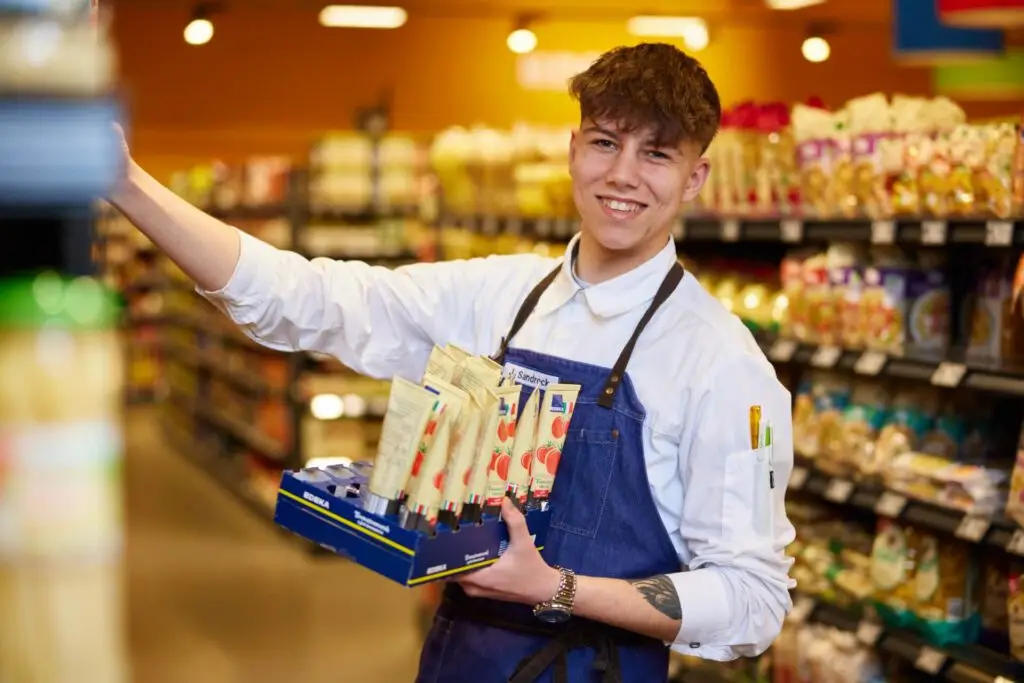 L&auml;chelnder junger Supermarktmitarbeiter in Sch&uuml;rze pr&auml;sentiert Tubenprodukte im Regalgang