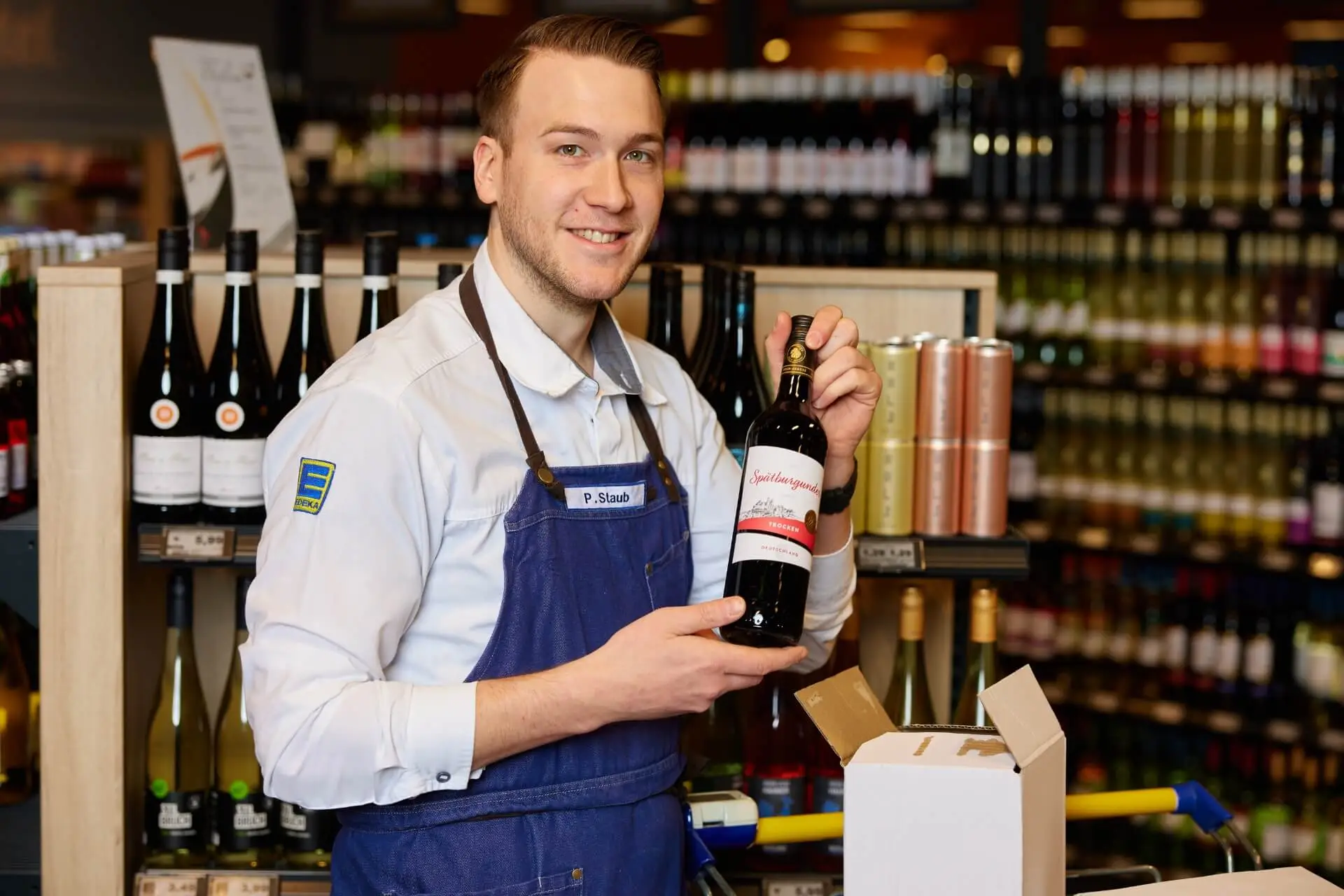 Weinverkauf: L&auml;chelnder Verk&auml;ufer pr&auml;sentiert Weinflasche vor Weinregal im Supermarkt, Verpackung sichtbar