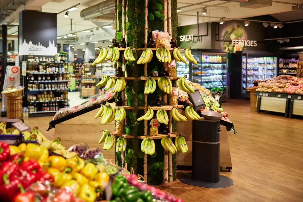 Supermarkt Obst & Gem&uuml;se: Bananen-Display an S&auml;ule, frische Paprika und weiteres Obst im Vordergrund