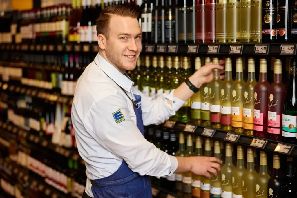 EDEKA-Mitarbeiter räumt Weinflaschen ins Regal im Supermarkt – gut sortiertes Weinangebot