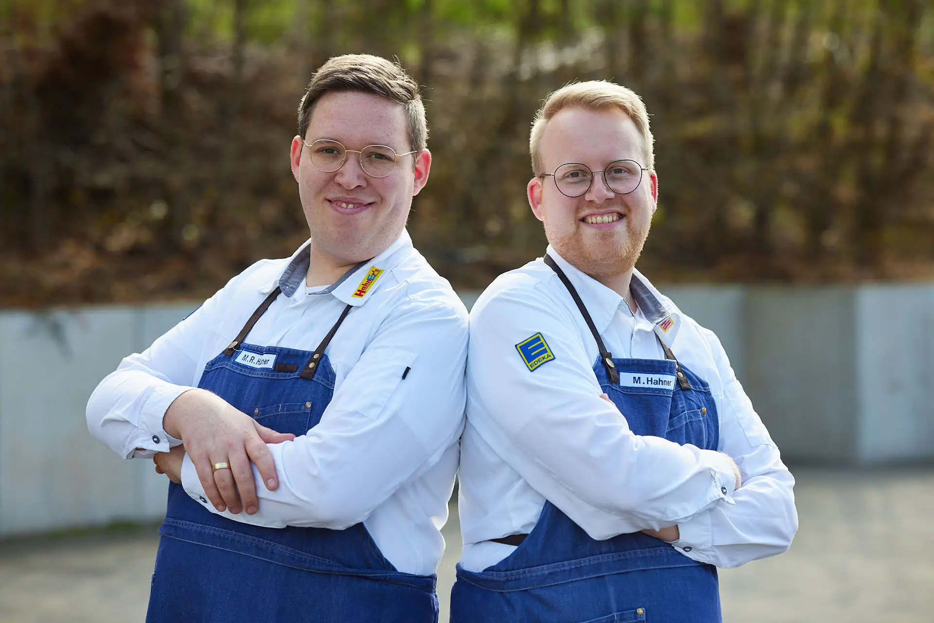 Zwei lächelnde EDEKA-Mitarbeiter im Team, weiße Hemden und blaue Schürzen, stehen Rücken an Rücken