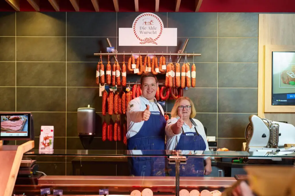 Wursttheke in Metzgerei mit aufgehängten Ahle-Wurscht-Salamis, zwei freundliche Mitarbeiter zeigen Daumen hoch